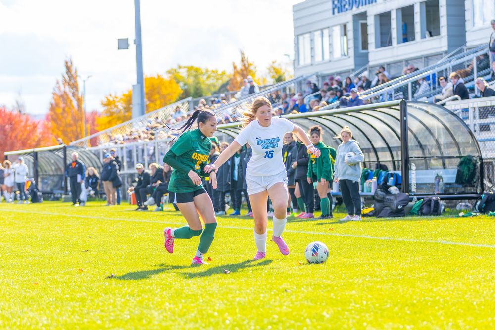 Soccer players compete for the ball during a match on a sunny day with spectator stands in the background.