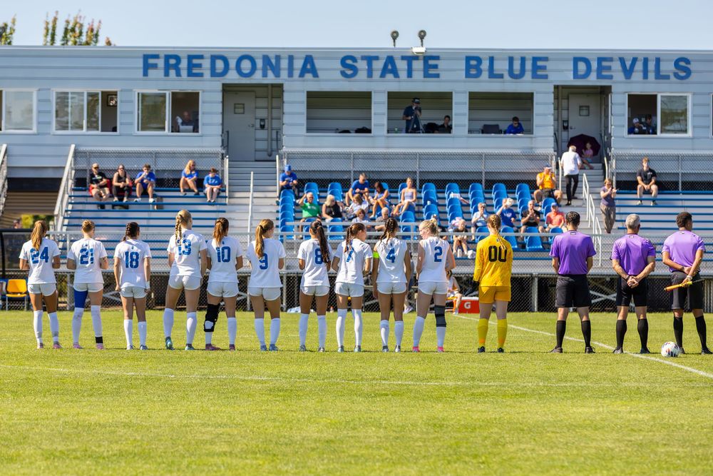 Soccer teams line up on the field before a match at Fredonia State Blue Devils stadium with spectators in the stands.