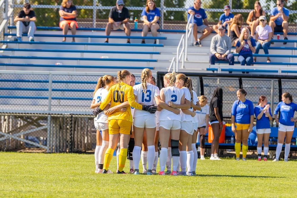 Soccer players sit on the sideline bench during a sunny match day while spectators watch from the bleachers.