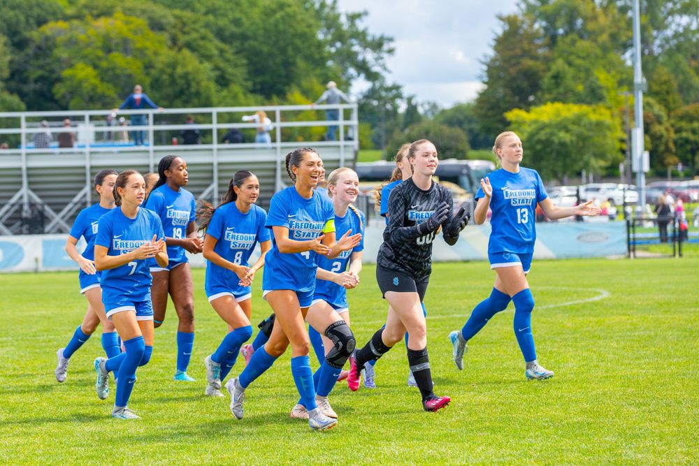 Soccer players in blue uniforms perform dynamic stretching exercises on a grass field during warmup drills.