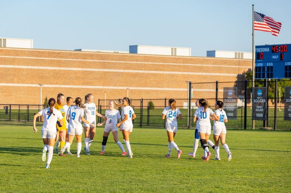 Soccer players in white uniforms walking across a grass field with an American flag visible in the background.