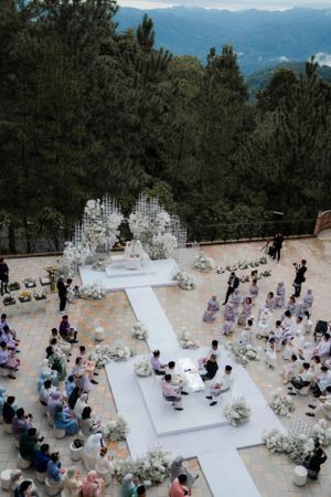 Aerial view of outdoor wedding ceremony with white cross-shaped aisle and floral arch in mountain setting.