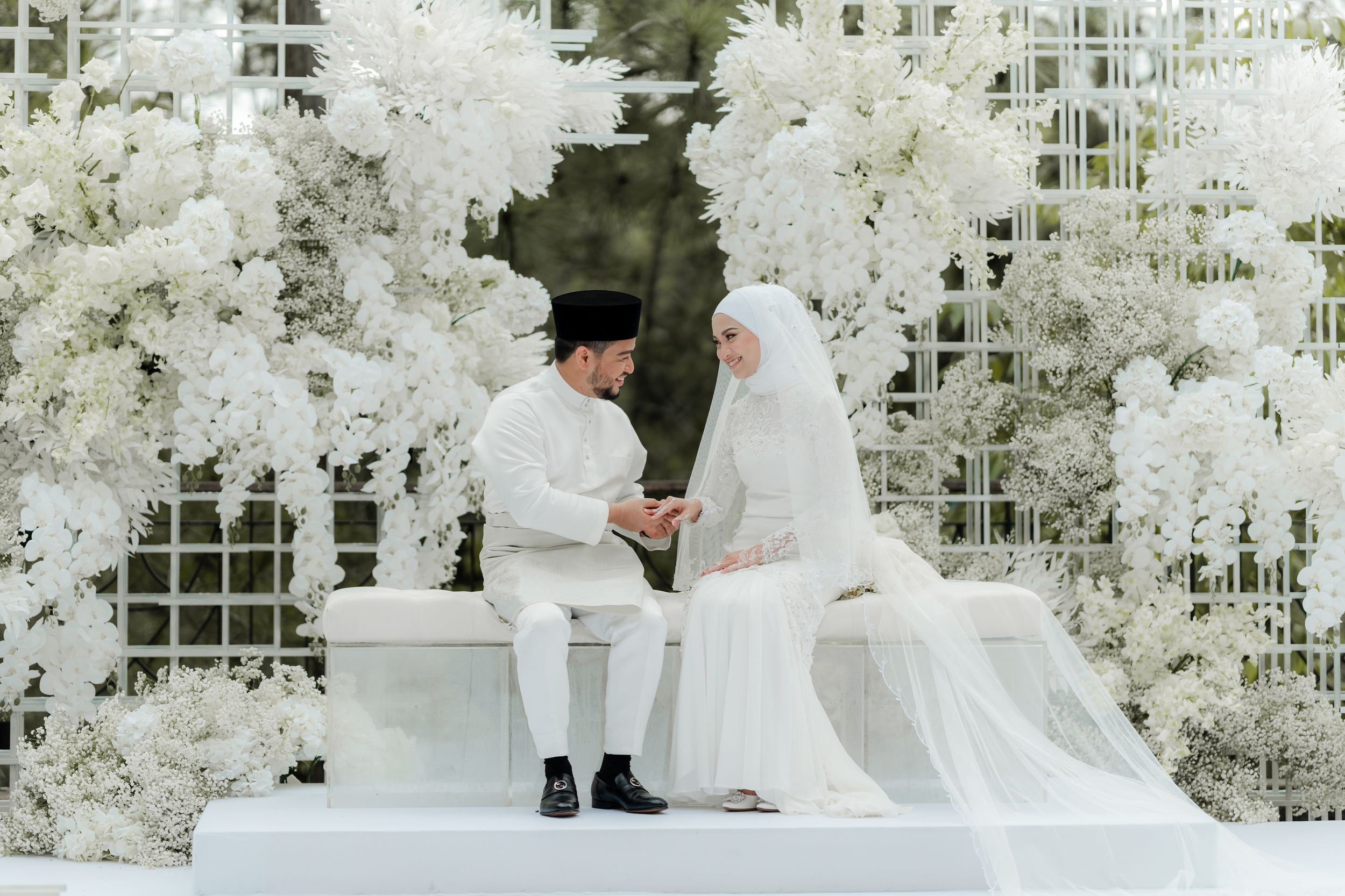 Wedding ceremony setup with couple seated on white bench surrounded by white floral arrangements.