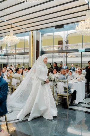A bride in white attire walks through an elegant reception hall with crystal chandeliers.