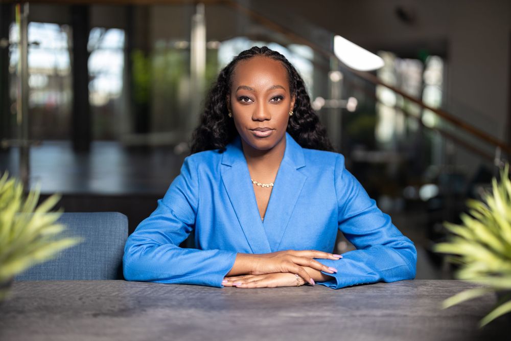 Professional portrait of woman in blue blazer seated at dark table with decorative plants.