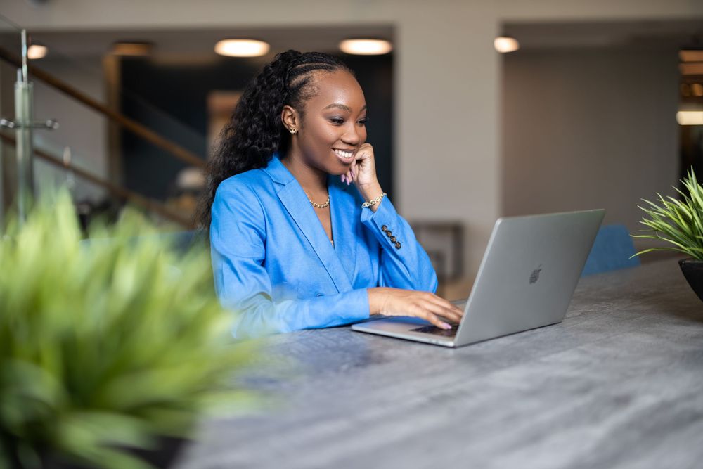 Professional working on laptop at modern desk with greenery visible in background.