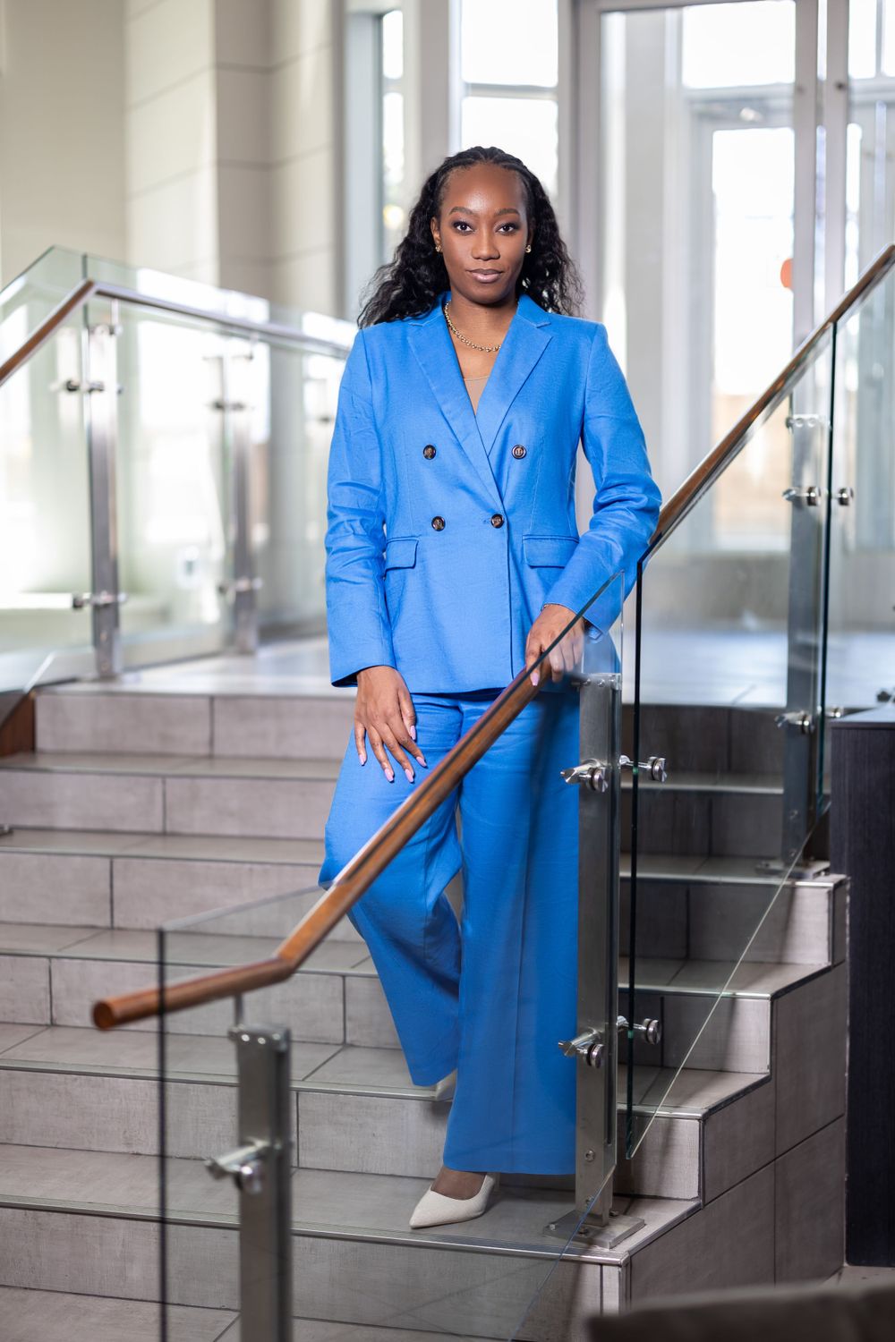 Business professional in bright blue suit poses elegantly on modern staircase with glass railings.