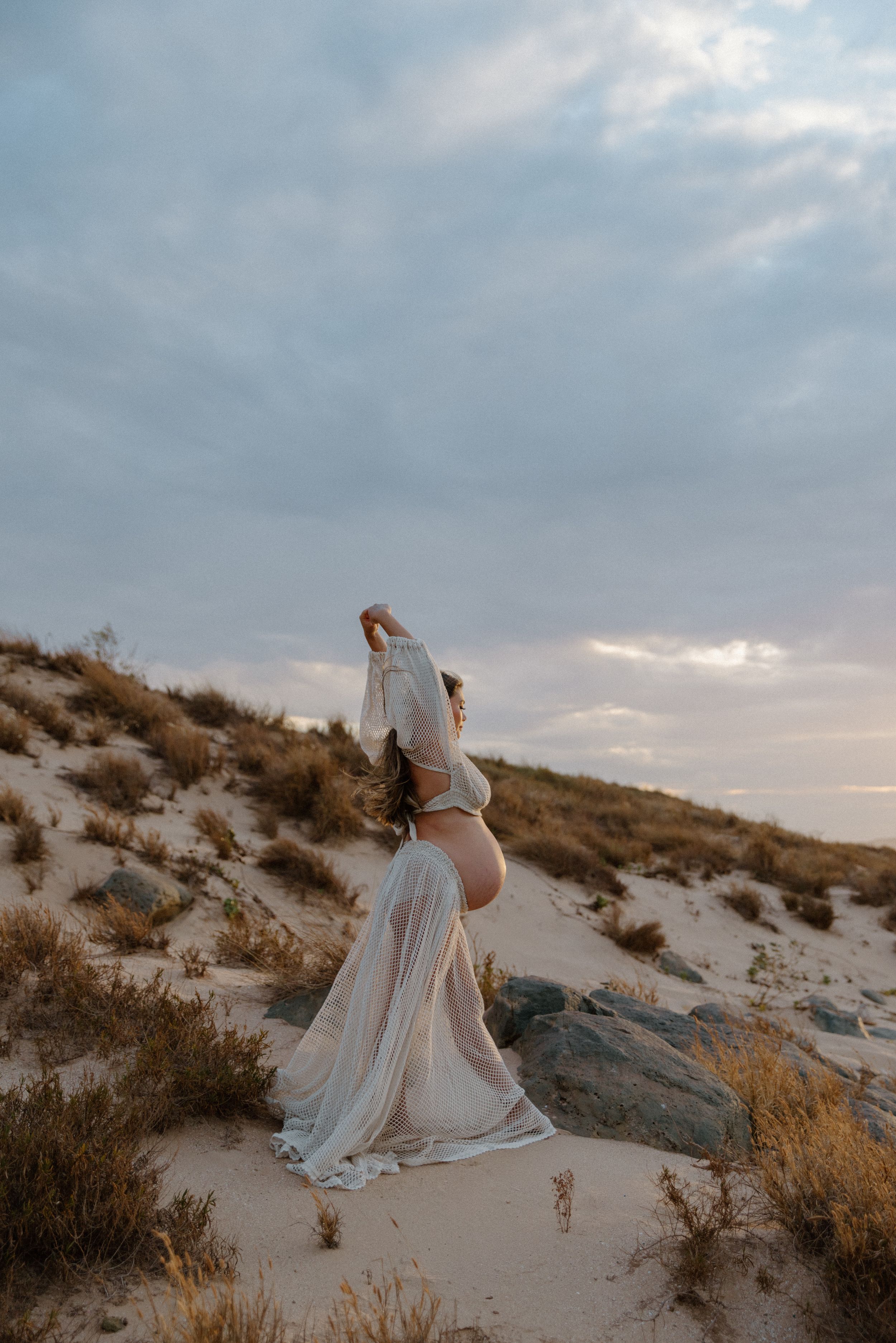 A person in a flowing white dress sits gracefully on sandy dunes during golden hour against a cloudy sky.