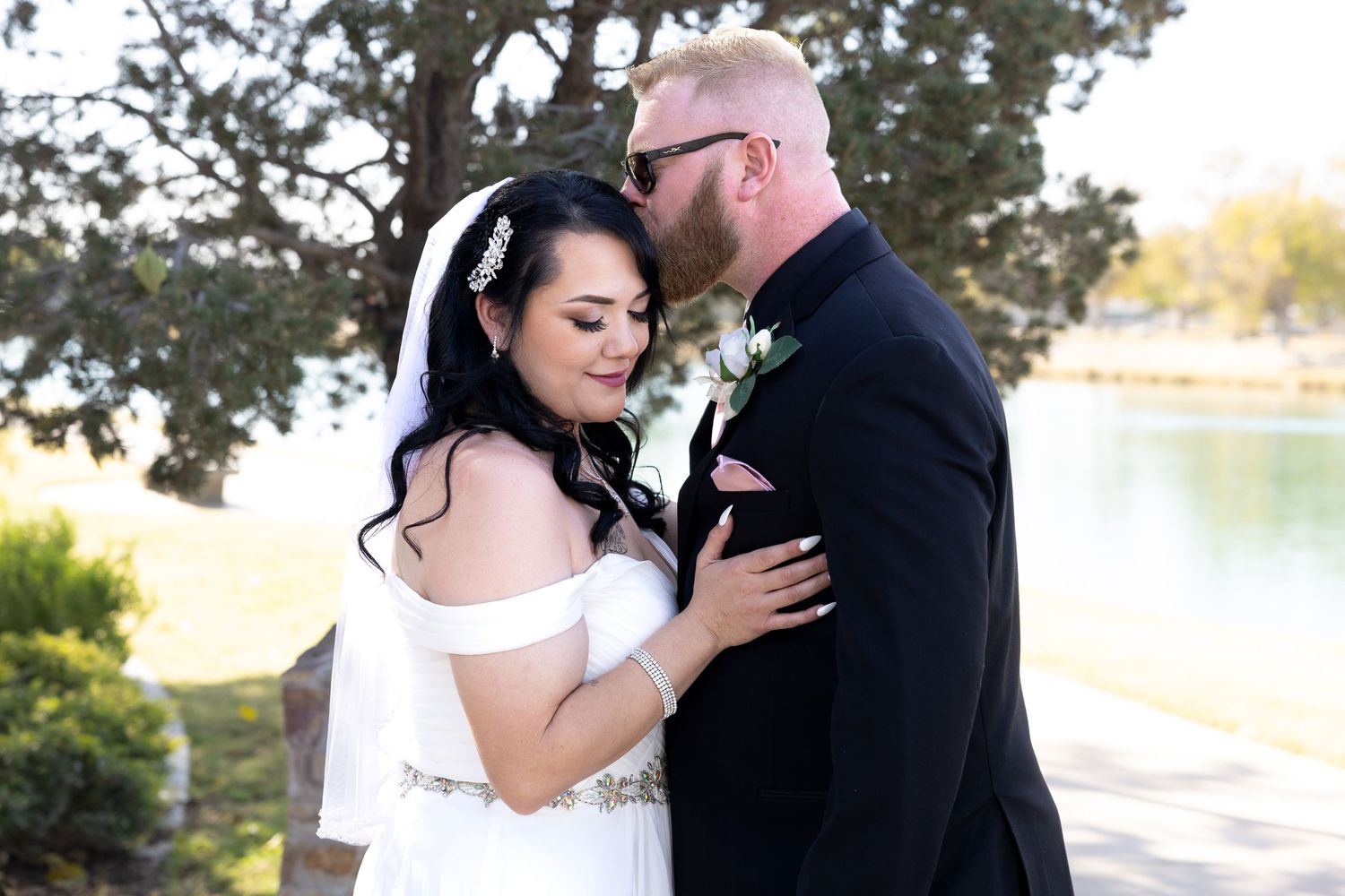 A bride and groom share an intimate moment together by a lake during their outdoor wedding photoshoot.