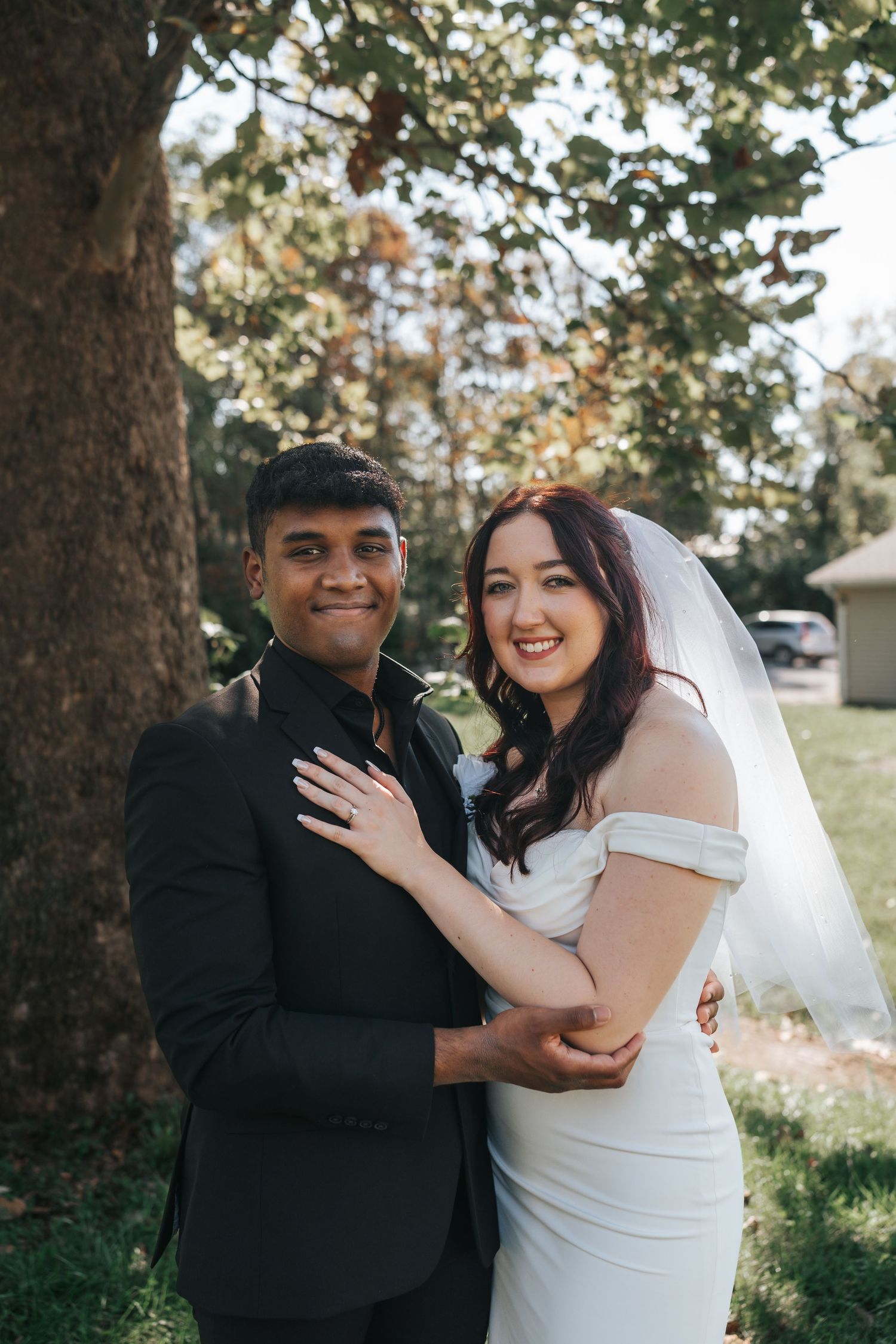 Romantic outdoor portrait of newlyweds in classic wedding attire beneath large tree.