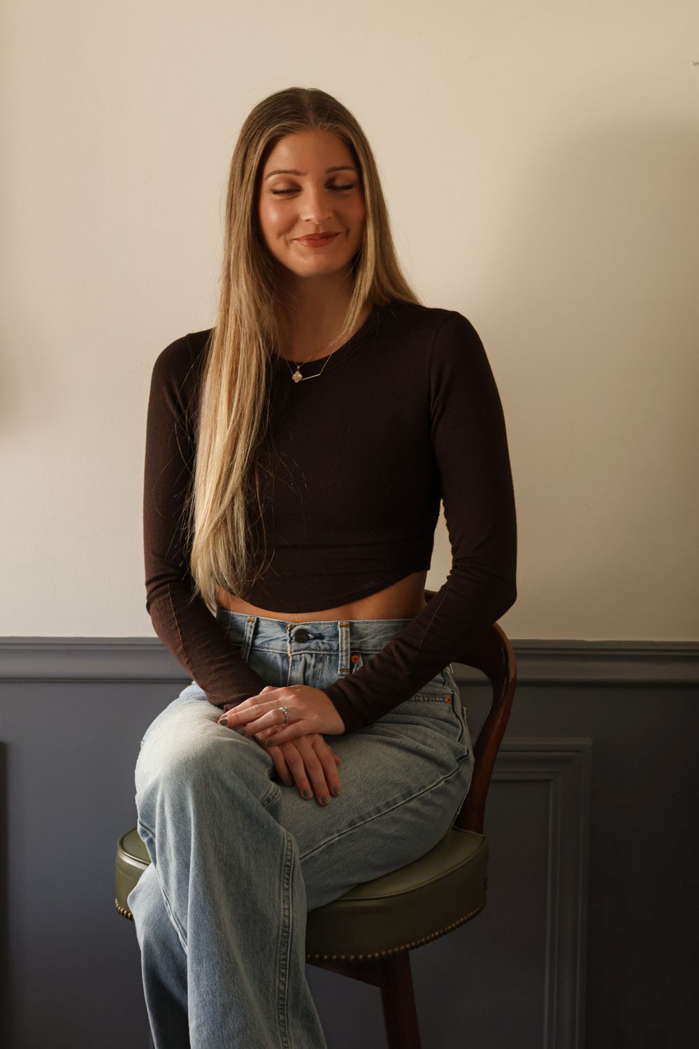 Person in dark crop top and light denim poses on chair against two-toned wall.