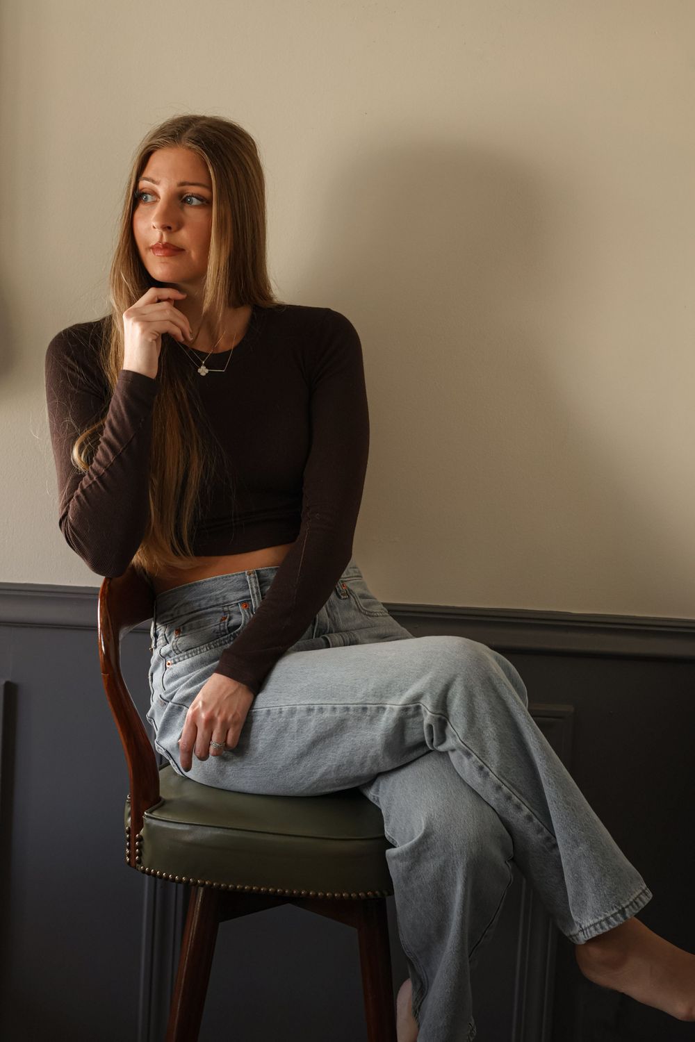 Person in black top and light jeans sitting casually on a chair against a neutral wall.