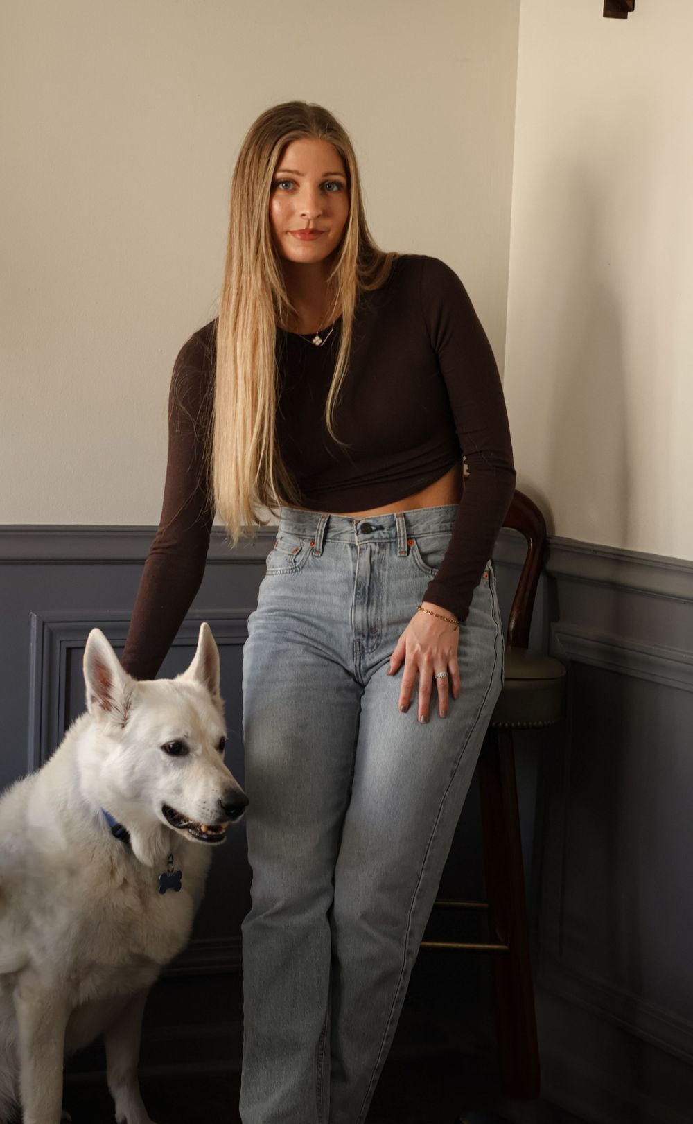 A white dog looking up at someone wearing casual attire against a grey wall.