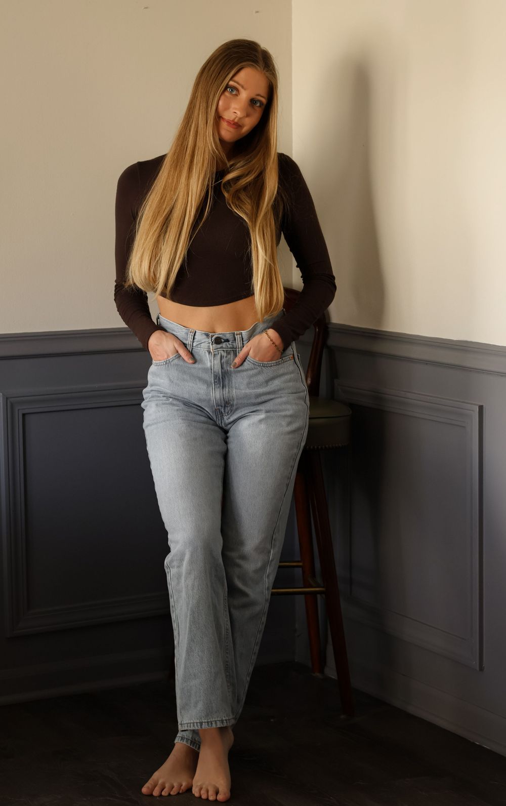 A relaxed pose on a wooden stool wearing jeans and a dark top against grey wainscoting.