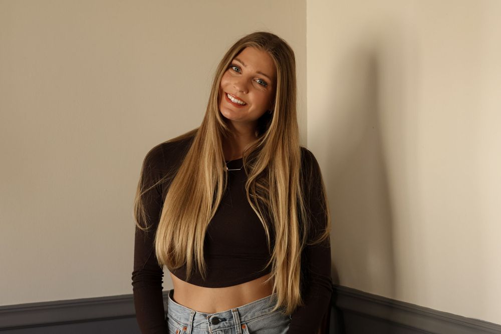 A casual indoor photoshoot featuring light denim jeans and a black crop top against a grey wall.