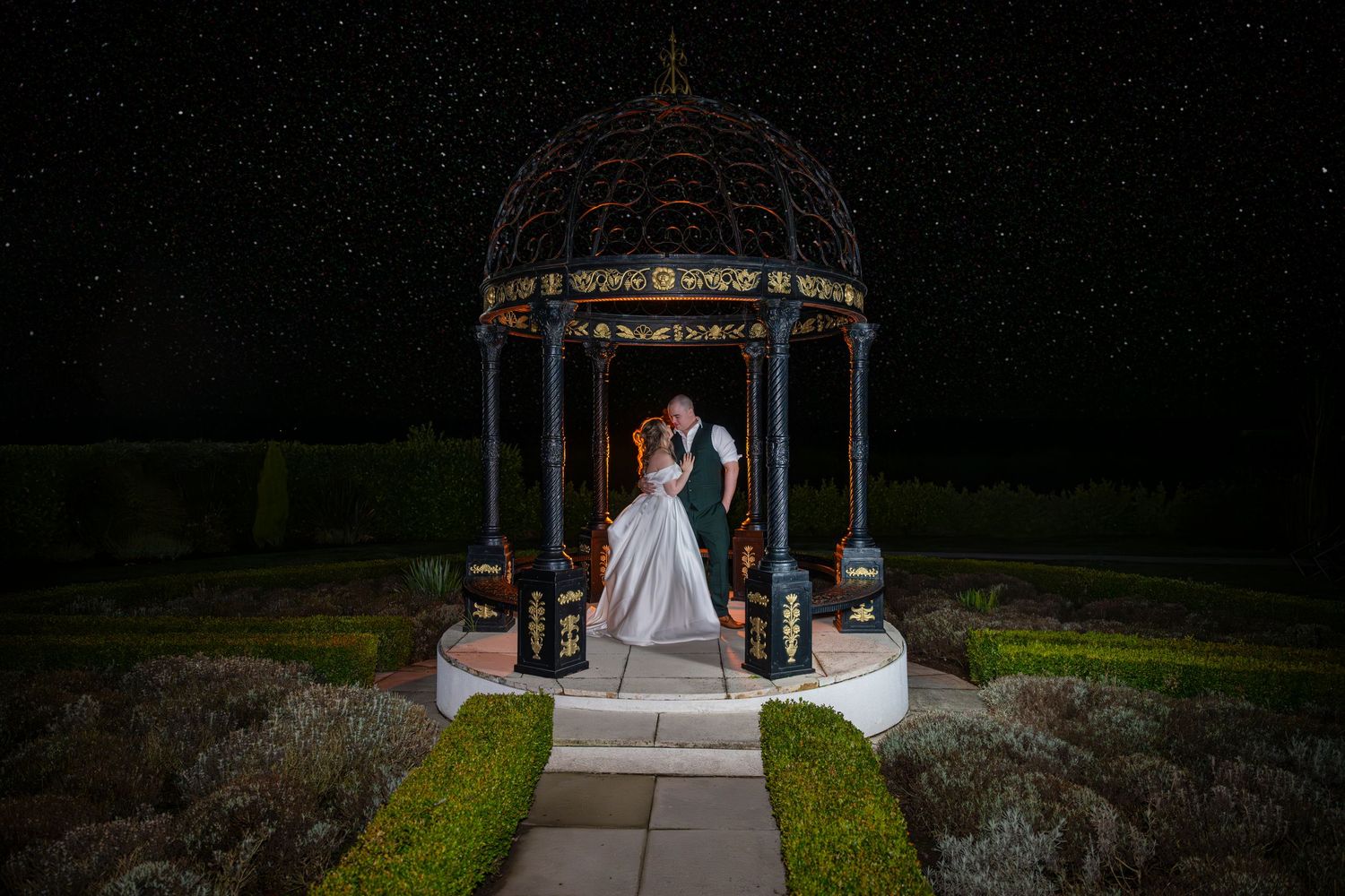 Bride and groom kissing under an ornate gazebo beneath a starry night sky in a formal garden.