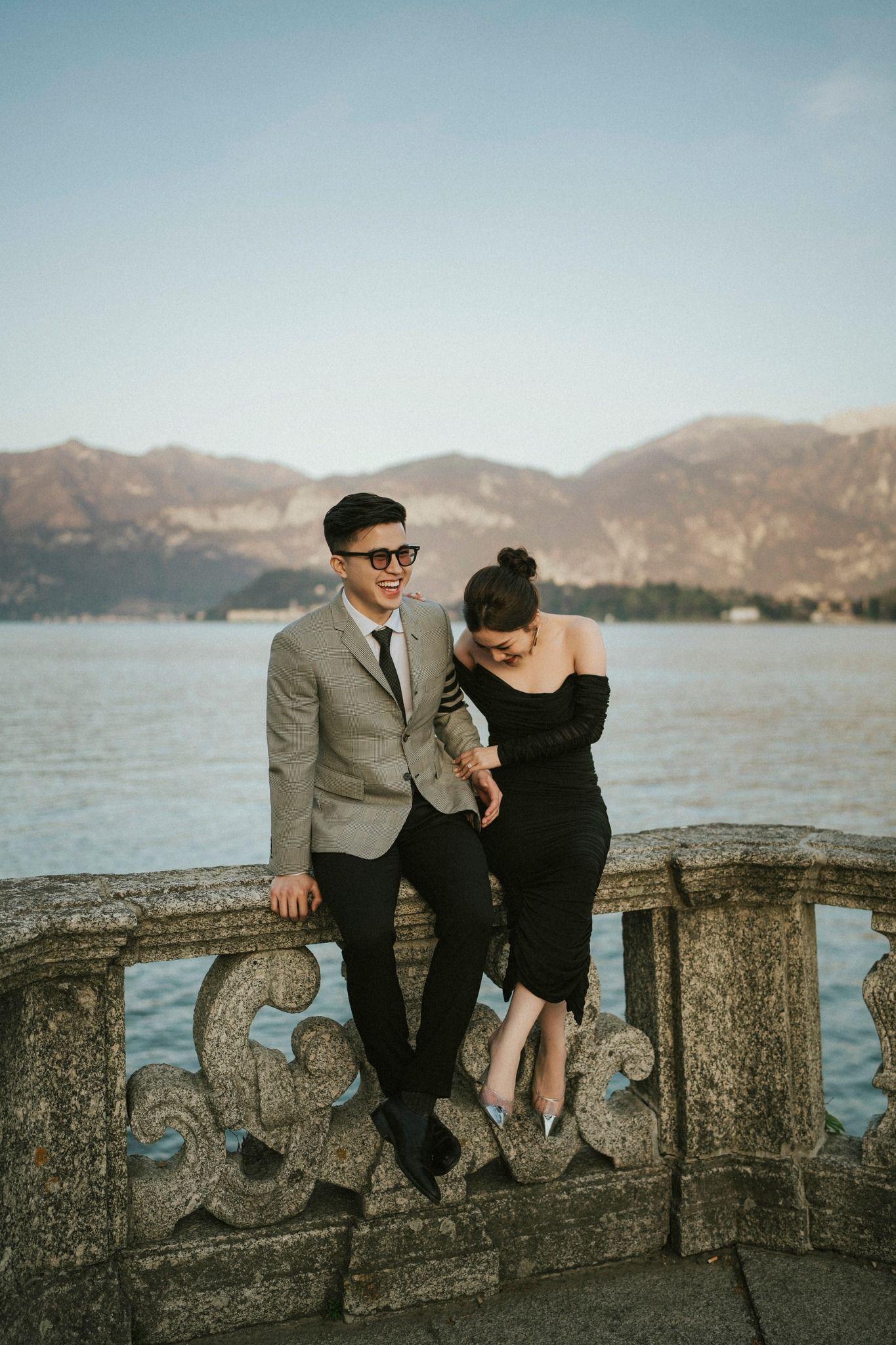 Stylish couple poses on stone balcony overlooking mountain lake landscape.