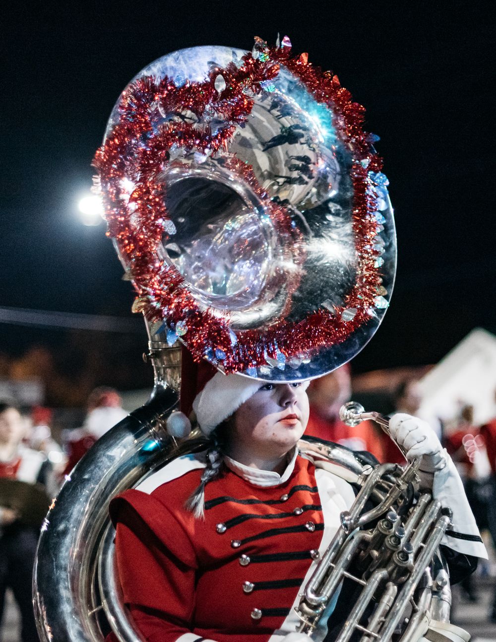 Marching band musicians play brass instruments decorated with red tinsel under stadium lights.