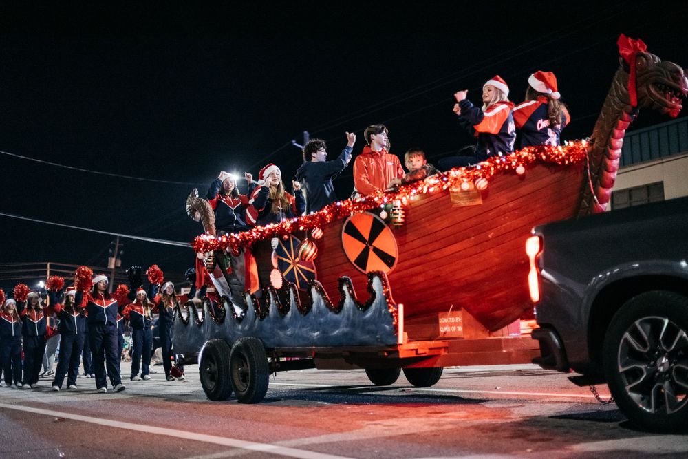 Parade participants lined up along parked vehicles with red holiday decorations at night.