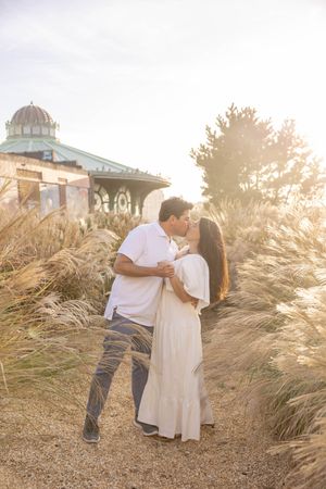 Couple shares a sunset kiss surrounded by tall golden grasses near a historic beachside pavilion in Asbury Park, NJ.