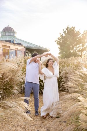 Joyful couple dances and twirls in golden sunlit field of tall grasses near a colorful pavilion in Asbury Park, NJ.