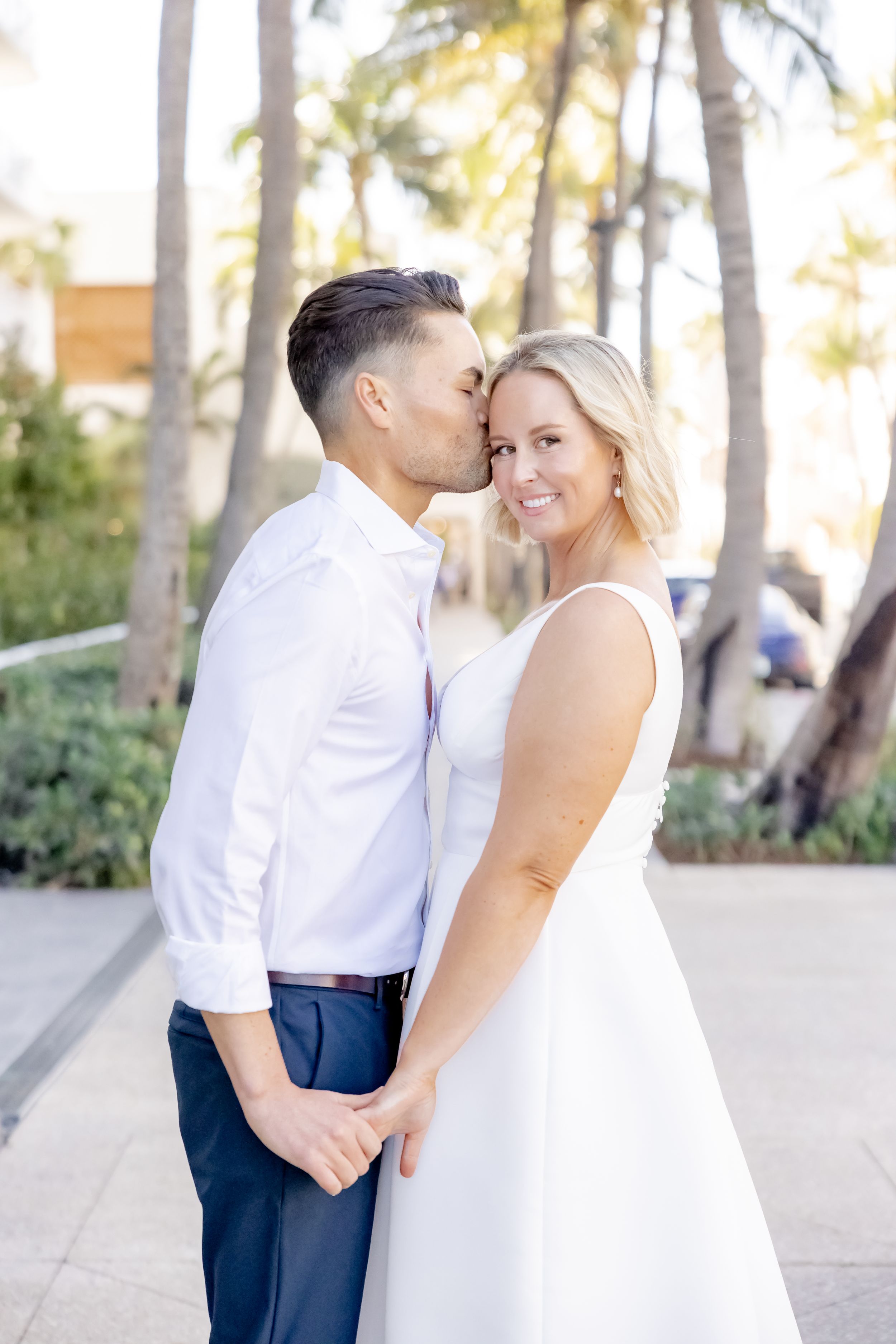 Man kisses woman on the cheek as she smiles at camera near tropical palm trees in Palm Beach, Florida.