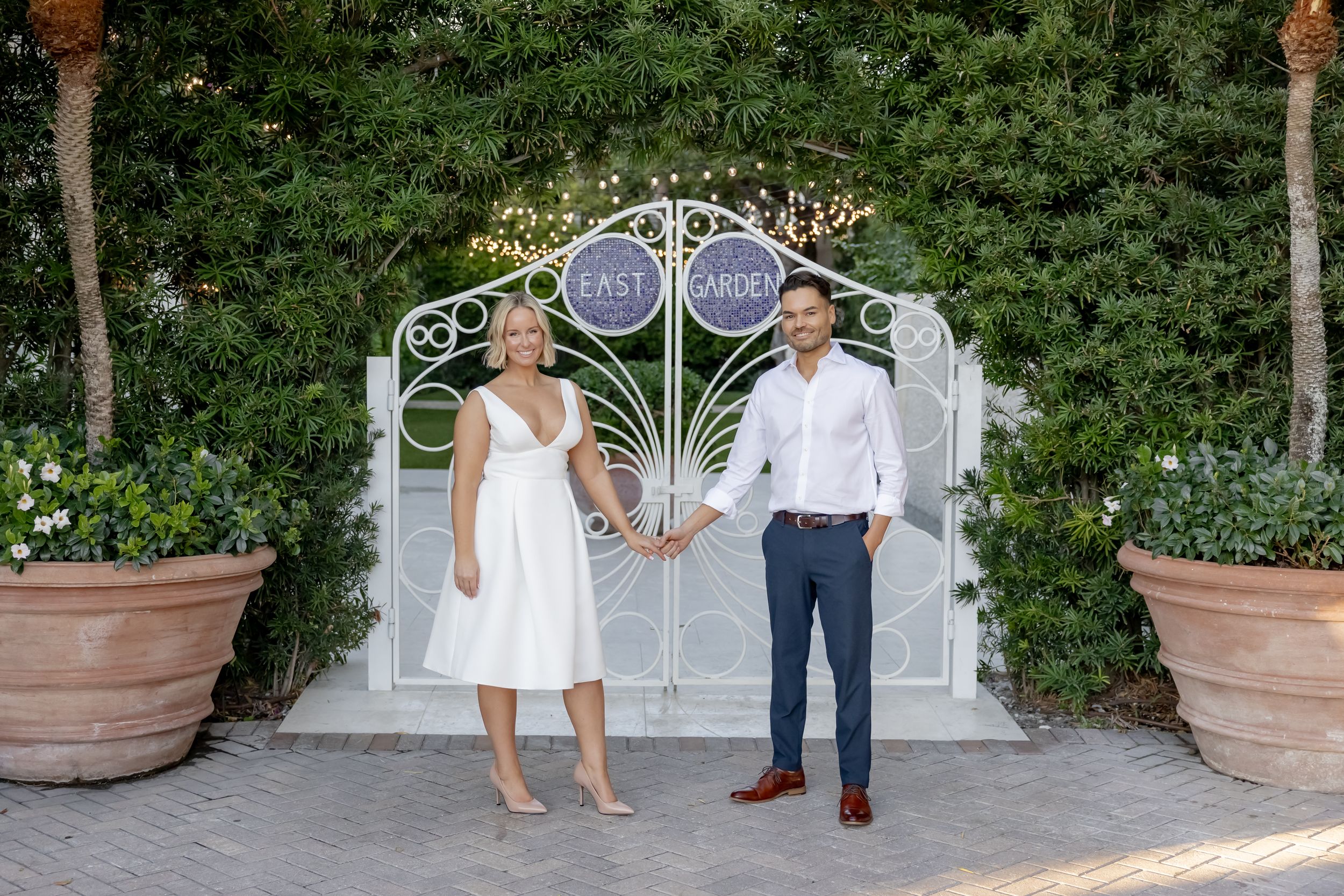 Couple holding hands in front of ornate white garden gate surrounded by lush greenery at elegant outdoor venue.