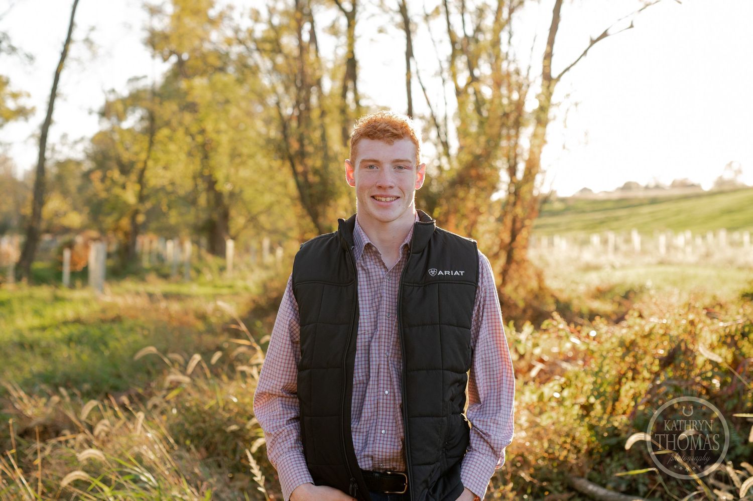 Outdoor portrait against golden trees wearing a black puffy vest and plaid button-down shirt.
