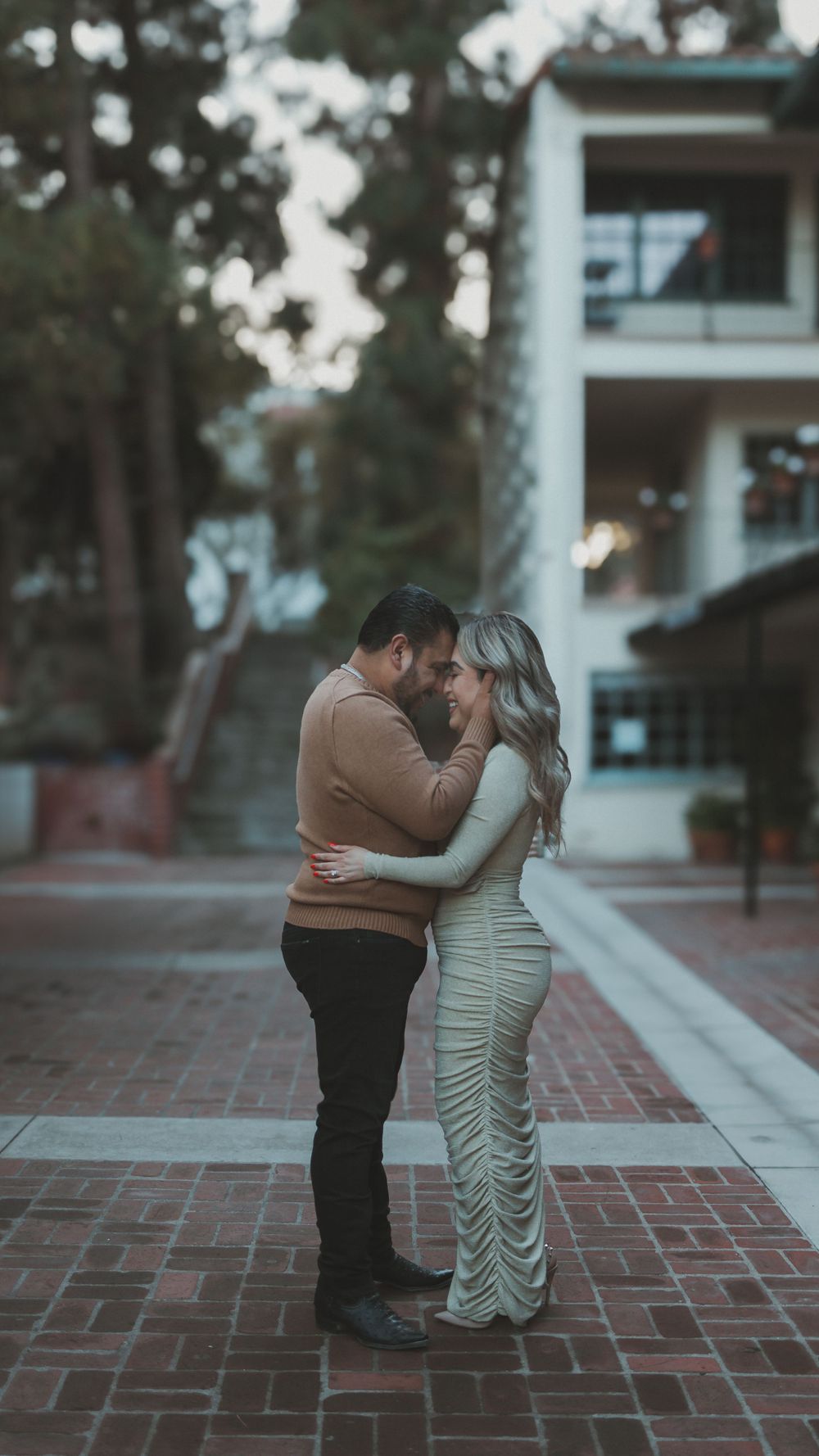 A romantic couple embrace on a brick pathway at dusk with modern architecture in the background.