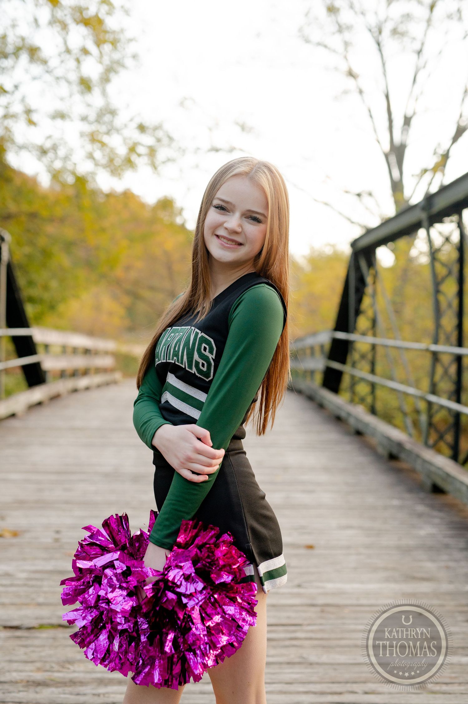Cheerleader in green uniform poses with bright pink pom poms on wooden bridge during autumn.
