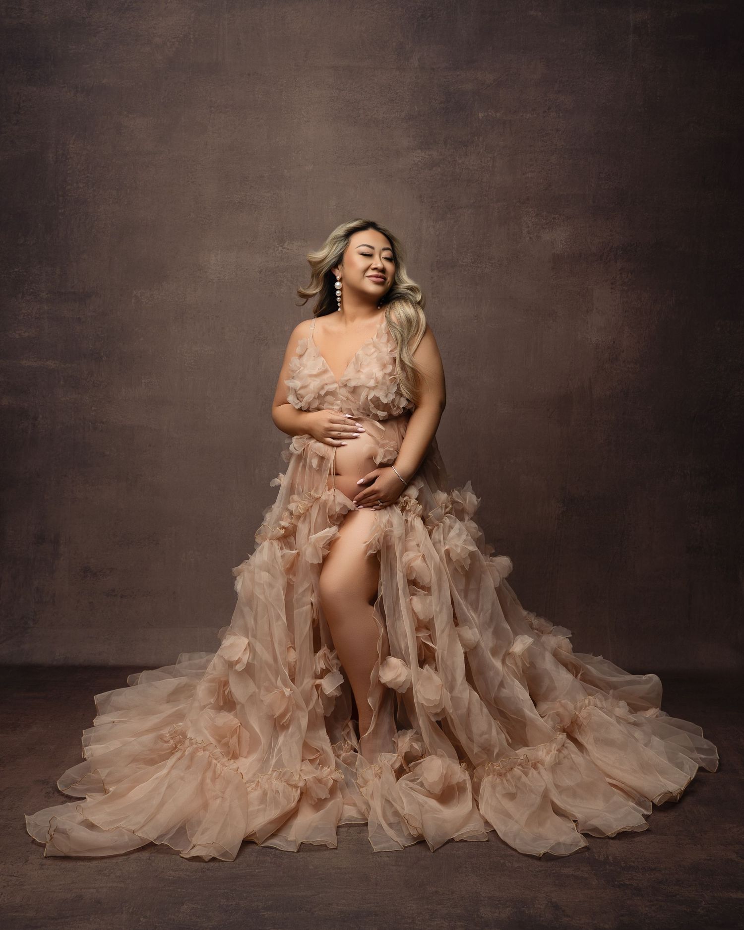 Elegant maternity photo series showing figure in flowing peachy-nude gown against dark textured backdrop creates dreamy effect.