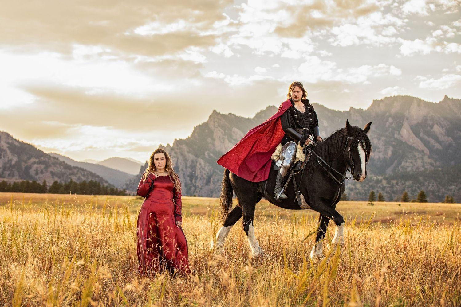 Two people in flowing red dresses pose dramatically in a mountain field at sunset.