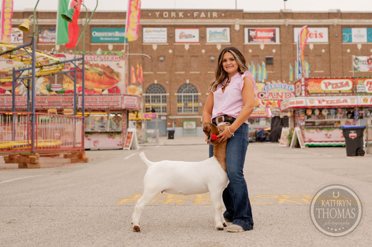 A goat stands with someone at a county fair with carnival rides and food stands in the background.