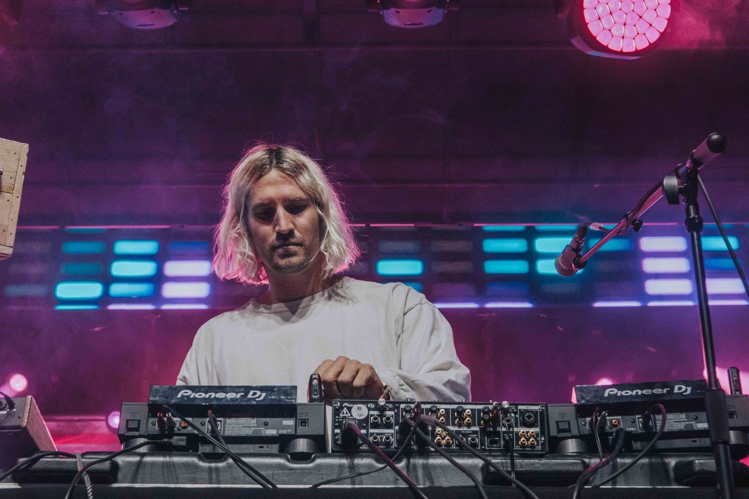 DJ performs at a nightclub with purple lighting and neon blue accents while standing at electronic equipment on stage.