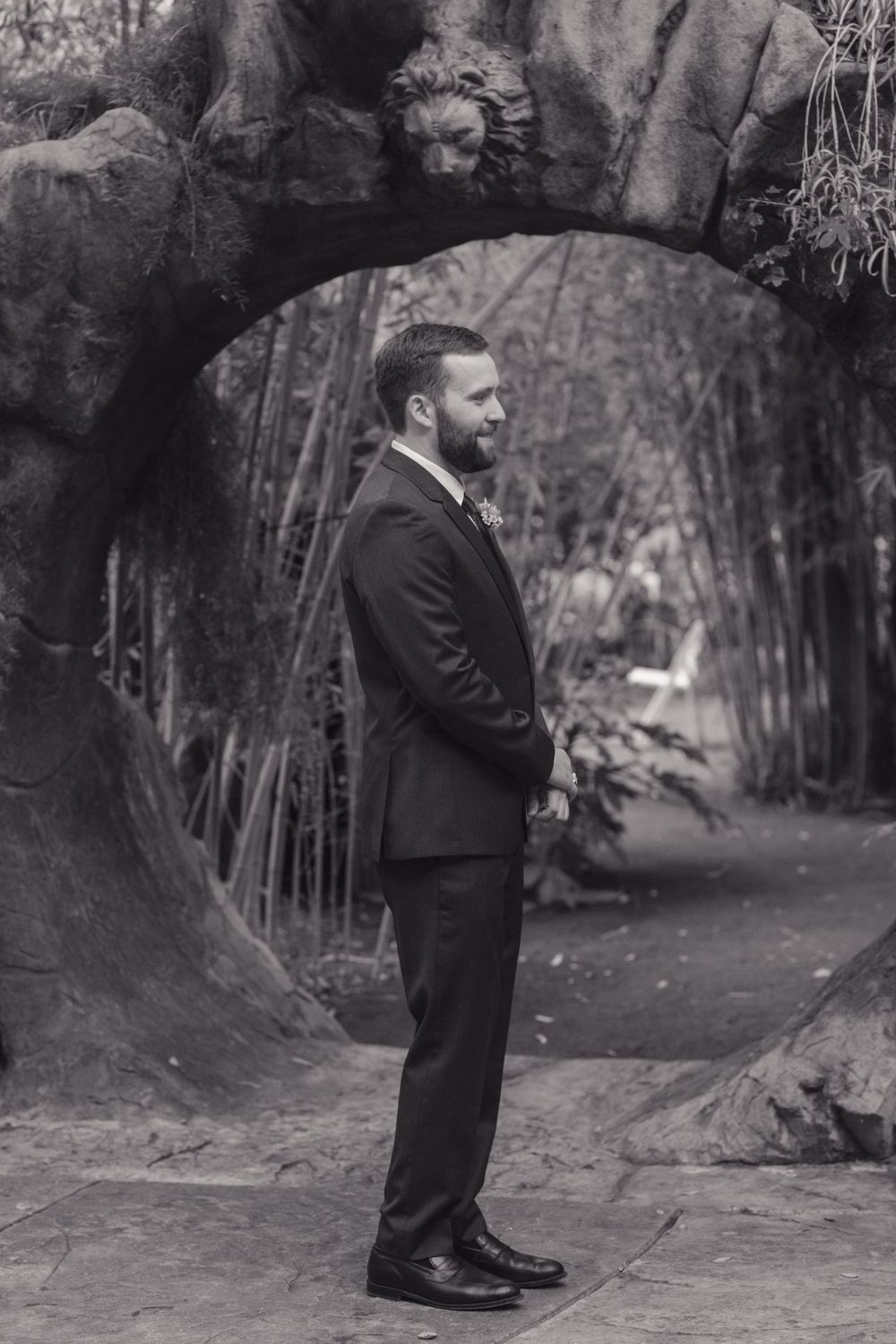A black and white photo of a groom in a formal suit standing under a stone archway in a garden setting.