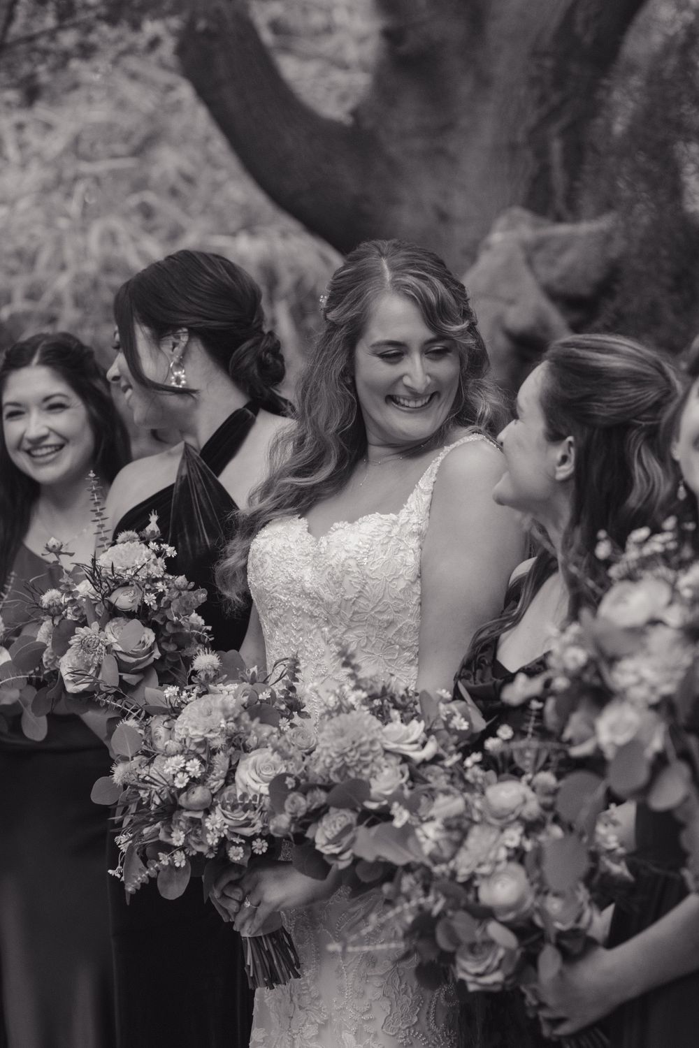 A bridal party in black dresses shares laughter while holding beautiful floral bouquets during an outdoor wedding ceremony.