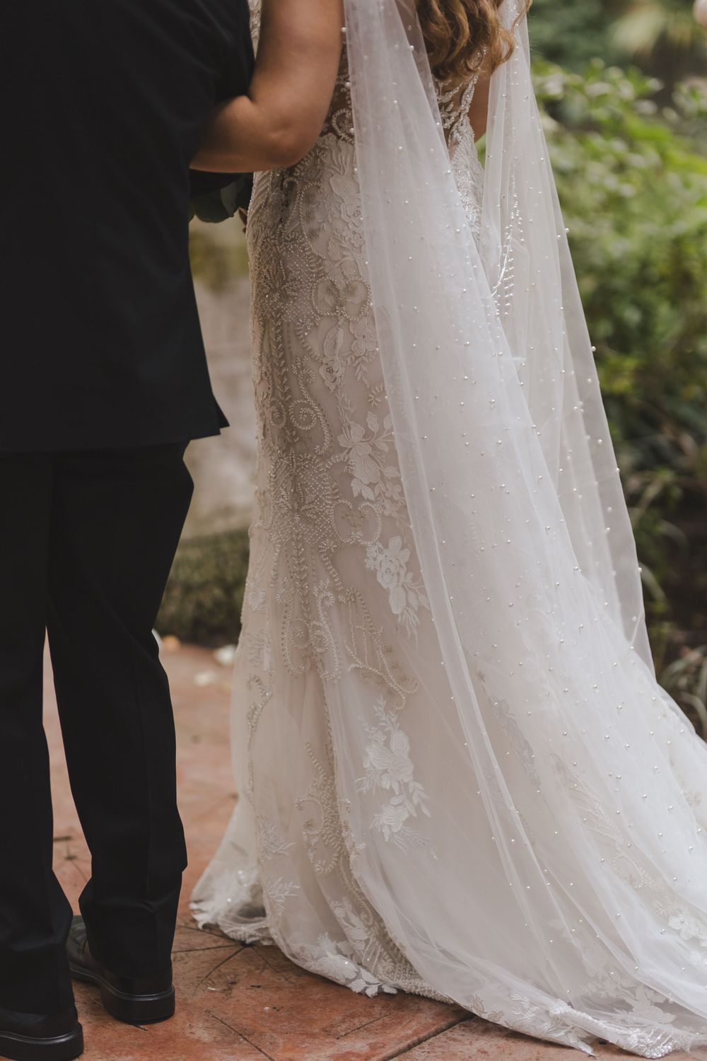 Close-up of elegant white lace wedding dress train and black suit at an outdoor garden wedding ceremony.