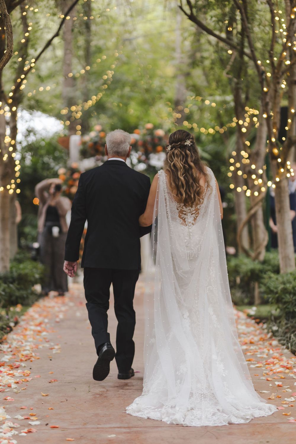 Bride and escort walking down an enchanted garden path lined with twinkling lights and autumn leaves.
