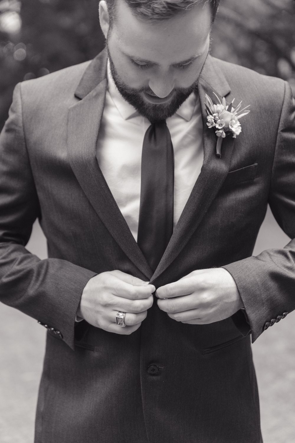 Black and white photo of a groom in a gray suit adjusting his jacket, wearing a boutonniere and wedding ring.
