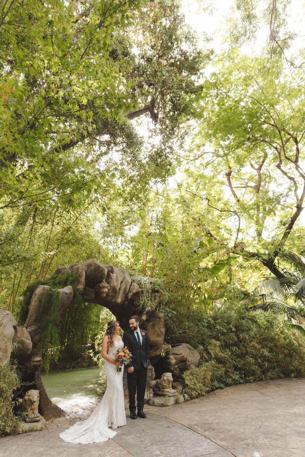 A romantic wedding couple poses under a stone archway surrounded by lush greenery in an outdoor garden setting.