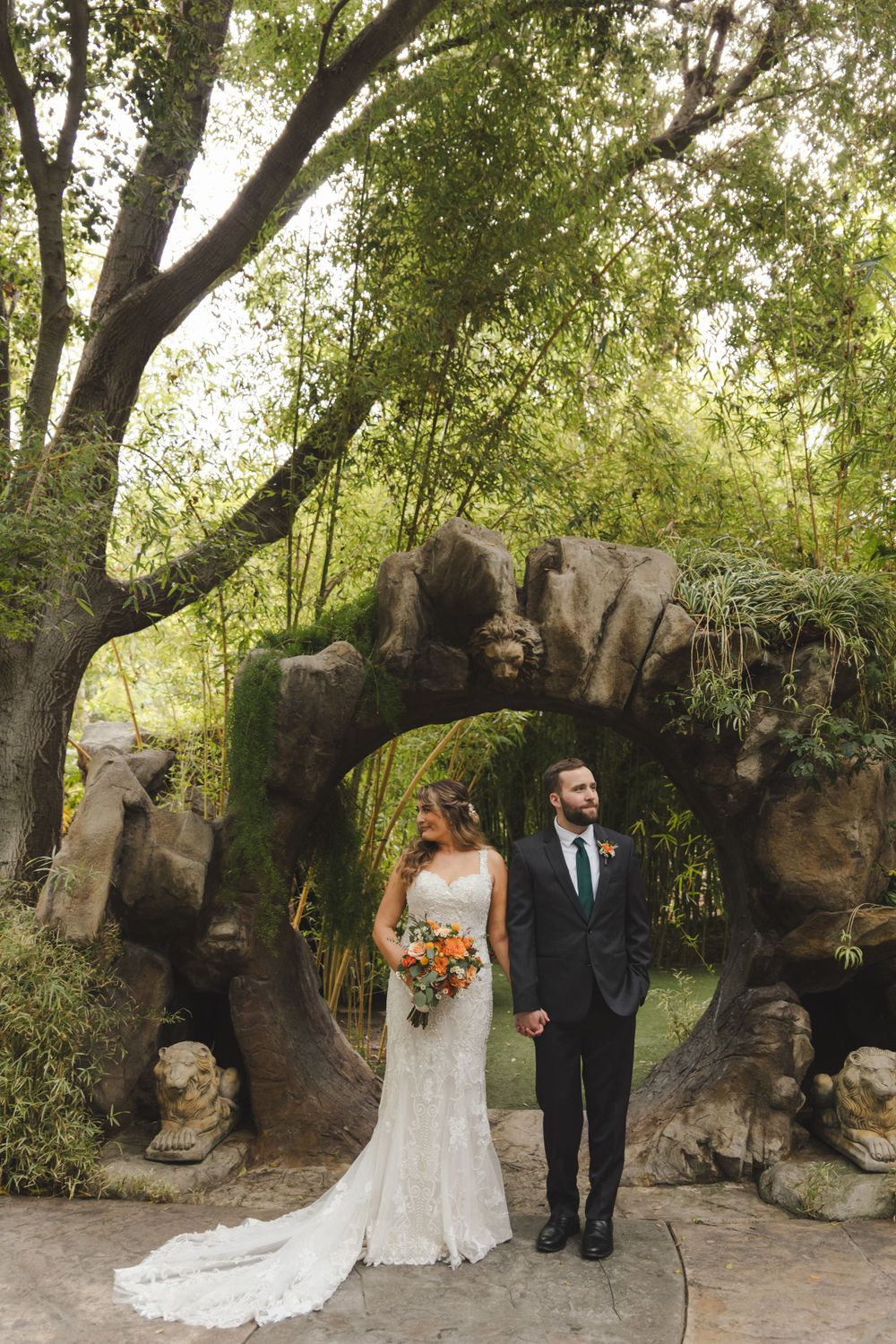 A couple stands under a stone archway surrounded by lush greenery in a romantic garden wedding setting.