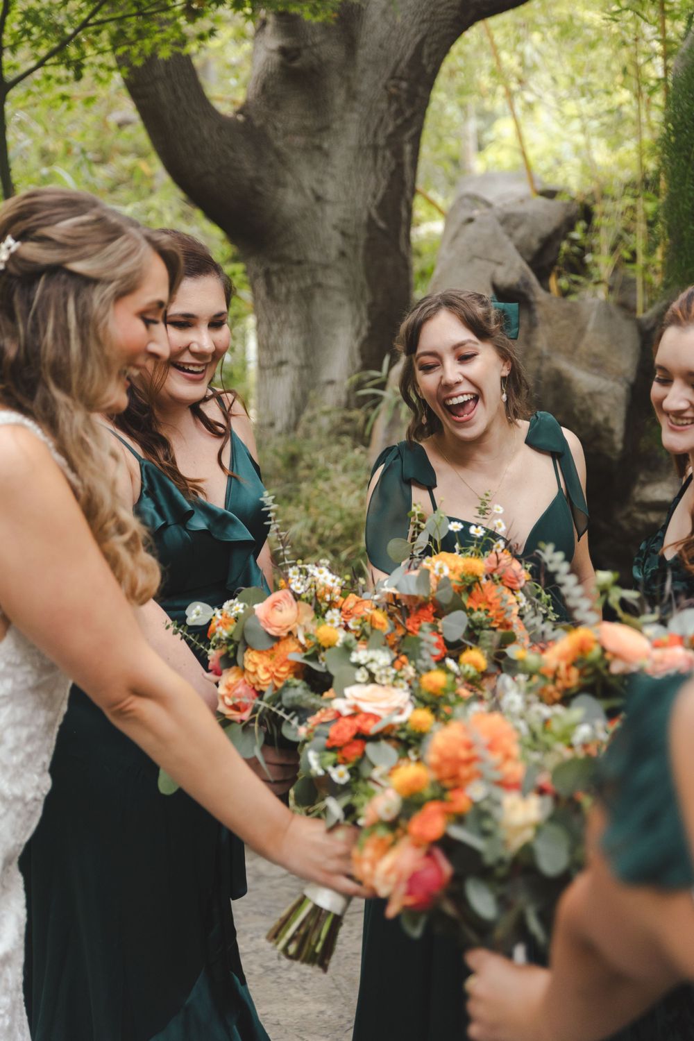 Bridesmaids in emerald green dresses share joyful laughter while holding vibrant orange and peach bouquets.