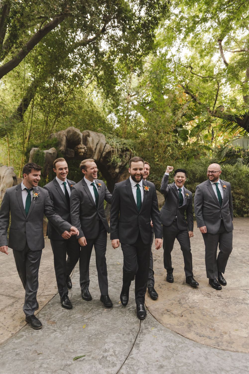 Groomsmen in dark suits walk together on a garden path under a canopy of green trees.
