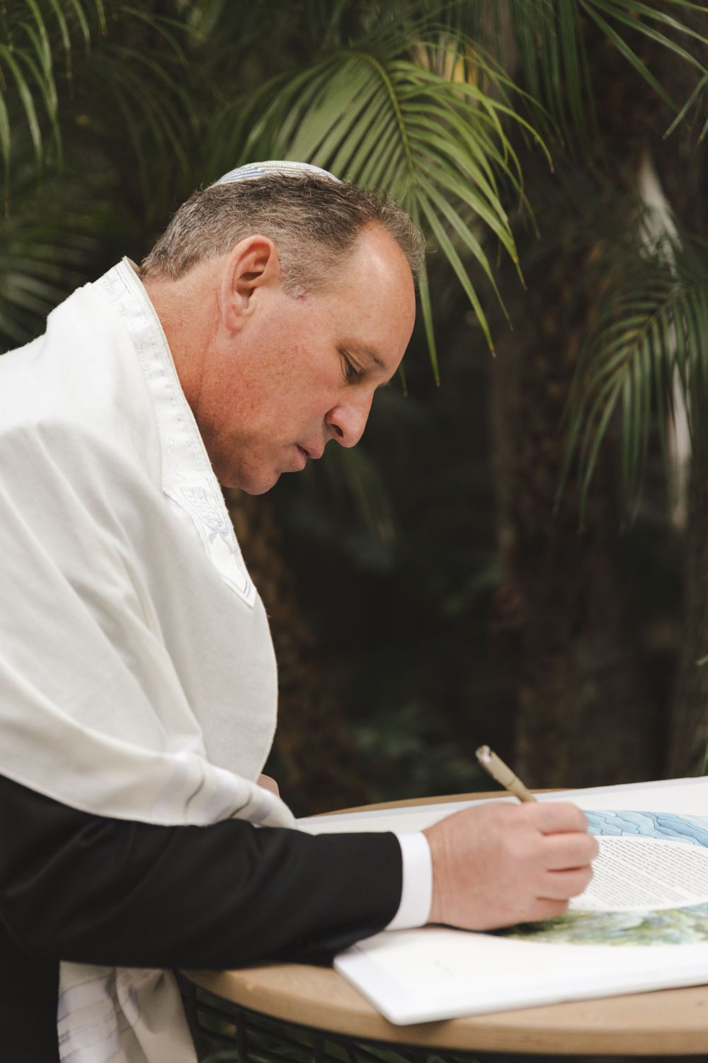 A focused man in white shirt and black suit writes in a notepad against a palm leaf background.