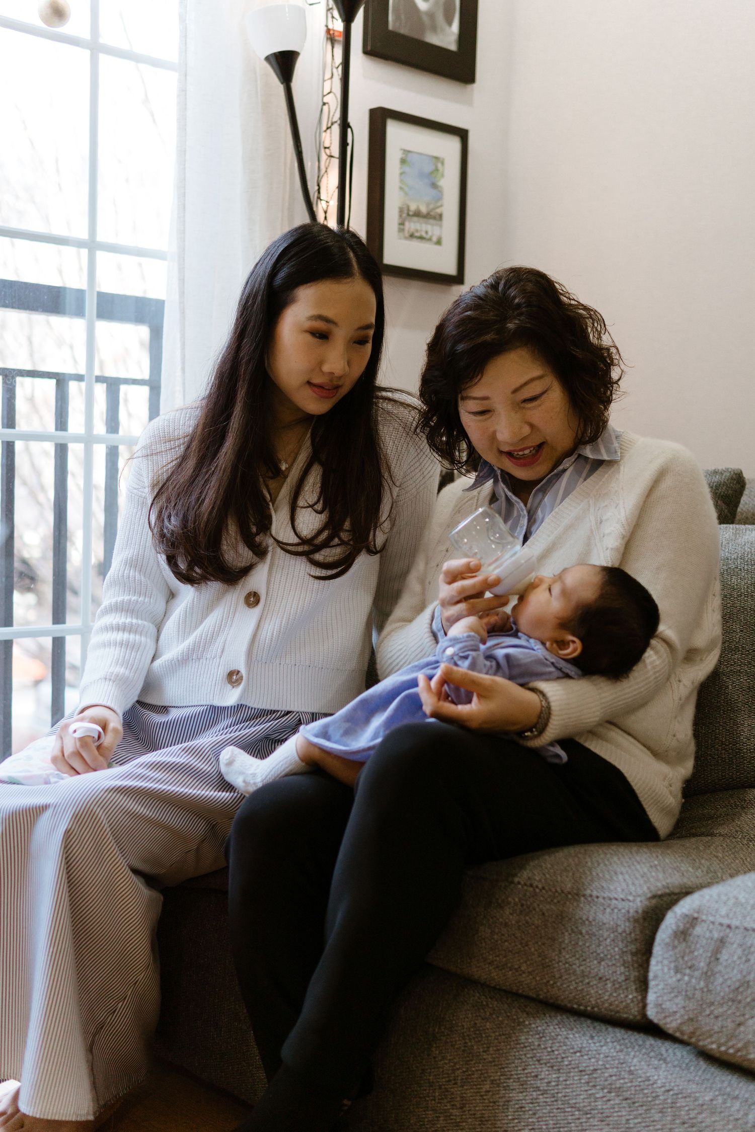 Three generations share a tender moment together on a cozy grey couch by a bright window during a newborn photoshoot in Philly.
