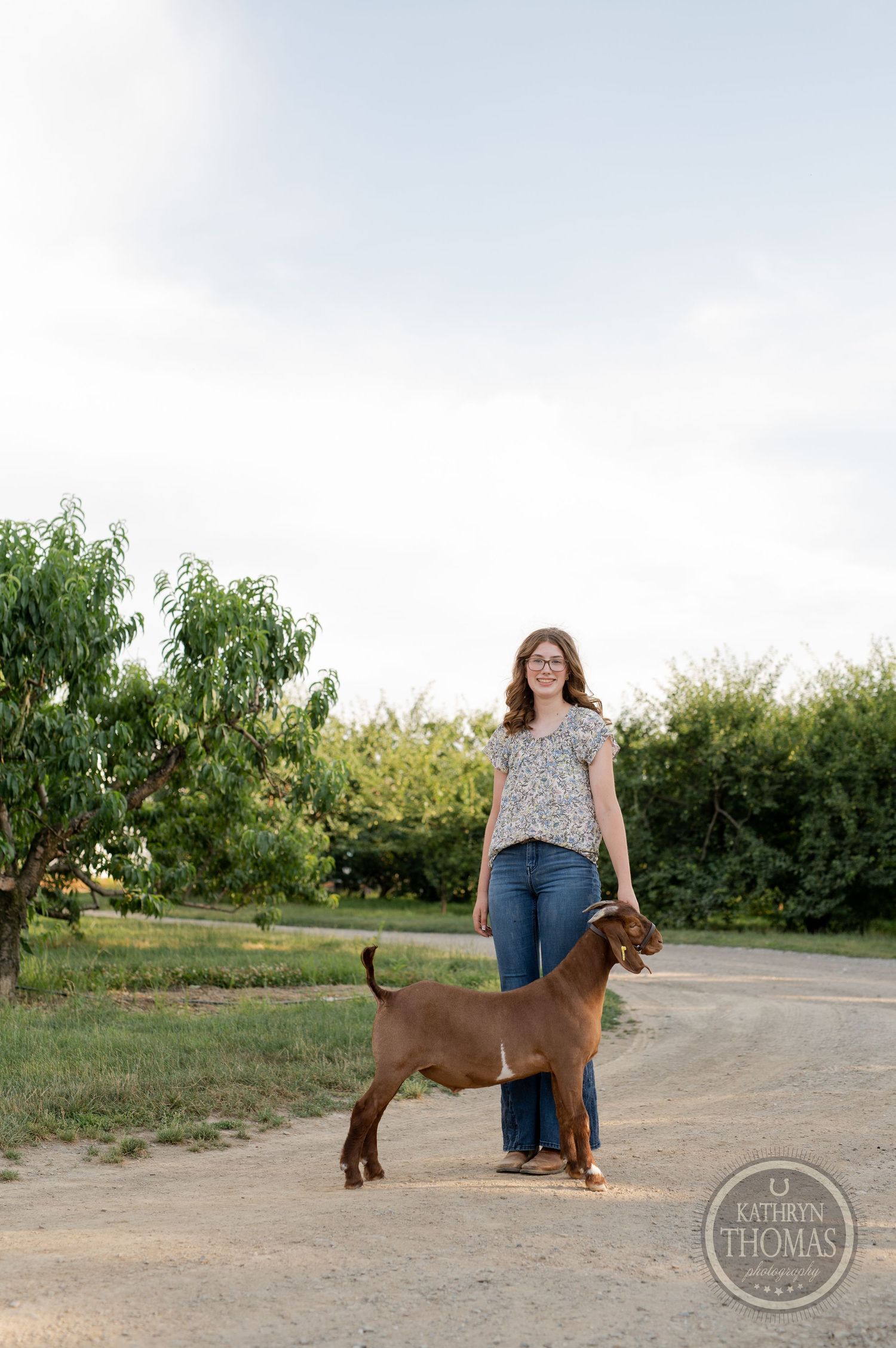 A brown goat poses with its handler on a rural pathway with trees and bushes in the background.