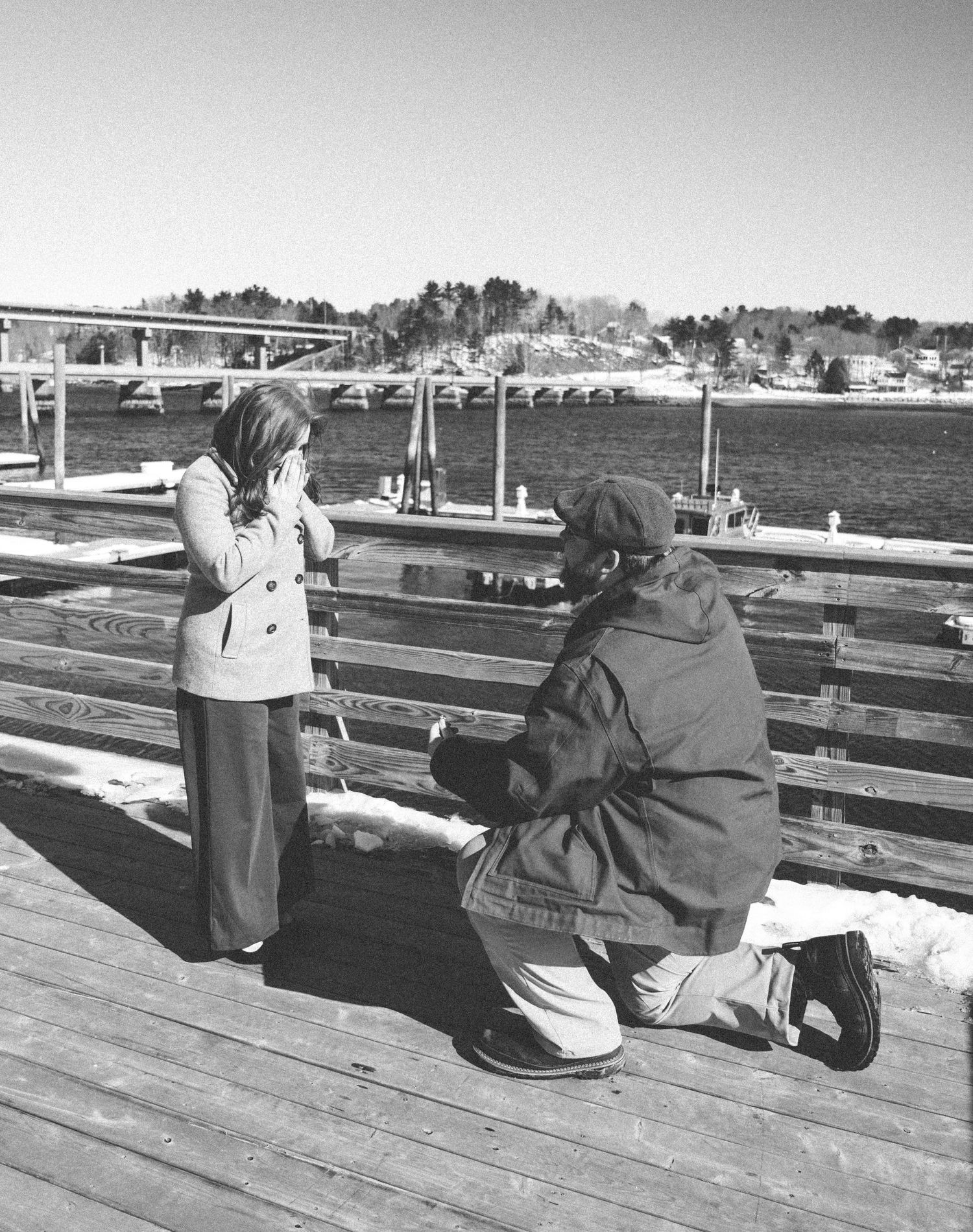 Proposal on a wooden dock overlooking a harbor with boats and trees in the background.
