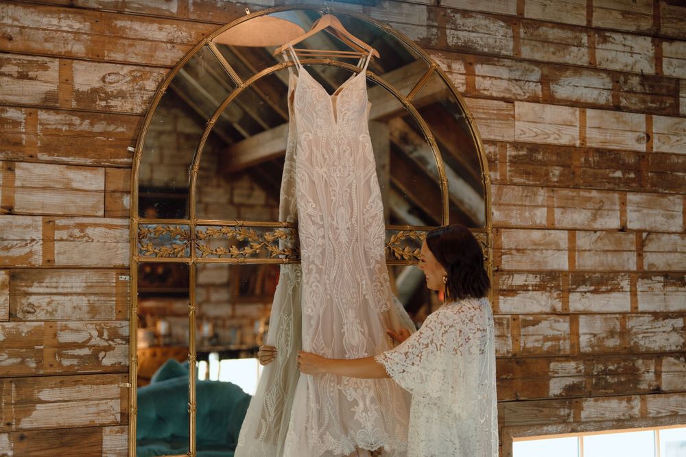 A white lace wedding dress hangs on a wooden hanger against a rustic brick wall with an arched window.