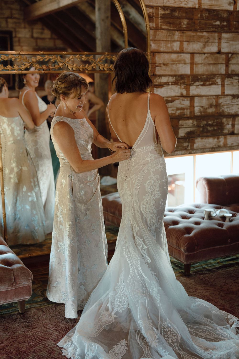 Two people in flowing white wedding dresses helping each other get ready in a rustic brick venue.