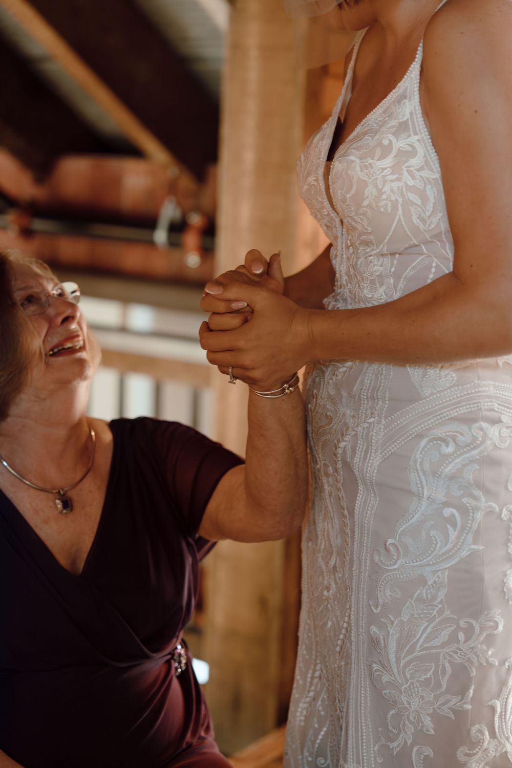 Two people share an emotional moment as one wearing a burgundy top helps adjust a beautiful white lace wedding dress.